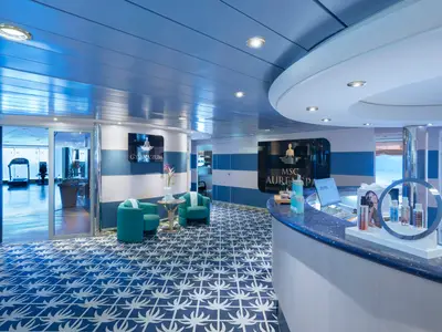 Ein stilvoller Empfangsbereich des MSC Aurea Spa an Bord der MSC Lirica, mit einer blauen Theke und zwei gruenen Sesseln. Durch Glastueren ist ein Fitnessraum mit Meerblick zu sehen, beleuchtet von warmem Licht.