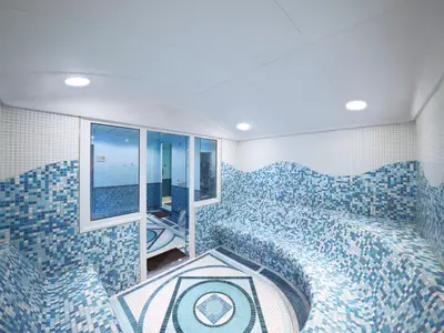 Blick in den Thermalbereich des MSC Aurea Spa an Bord der MSC Lirica. Der Raum ist mit blauen und weißen Mosaikfliesen gestaltet, inklusive geschwungener Sitzbänke und dem Boden. Links führen Glastüren in weitere Räume. Deckenleuchten erhellen den modernen Raum.