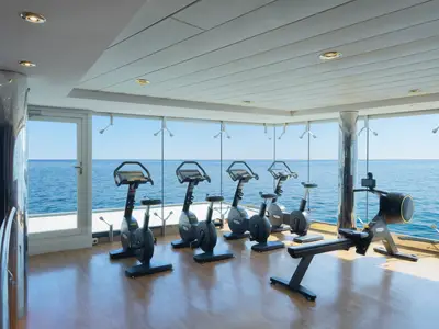 Ein moderner Fitnessraum an Bord der MSC Lirica mit mehreren Trimmrädern und einem Rudergerät. Große Fenster bieten einen weiten Blick auf das offene Meer.