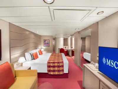 Helle Junior Suite Aurea an Bord der MSC Lirica mit grossem Doppelbett, orangeroter Bettdecke und Kissen. Links ein gelbes Sofa, rechts ein Schminktisch mit rotem Stuhl und Fernseher.