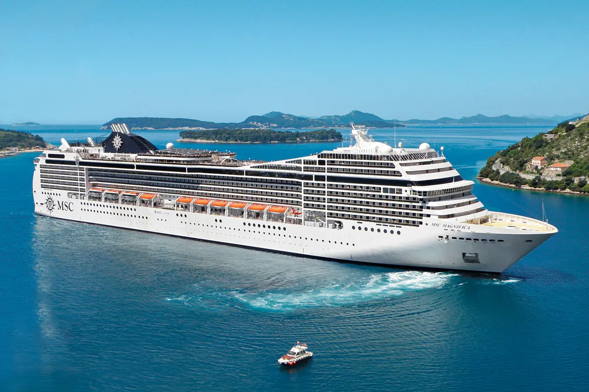 Die MSC Magnifica, ein großes weißes Kreuzfahrtschiff, fährt majestätisch durch blaues Meerwasser. Im Hintergrund sind grüne Inseln und eine mediterrane Küstenlinie mit Häusern zu sehen. Ein kleineres Boot folgt der MSC Magnifica.