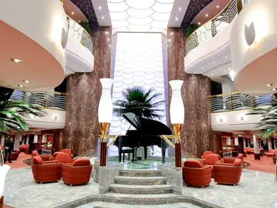Das elegante Atrium der MSC Magnifica. Im Zentrum steht ein schwarzer Flügel, umgeben von roten Sesseln. Der mehrstöckige Raum besticht durch marmorierte Wände und geschwungene Balkone.