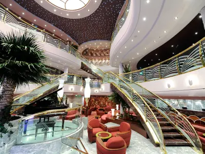 Das prachtvolle Atrium der MSC Magnifica zeigt geschwungene Treppen mit goldenen Gelaendern, ein Piano, Lounge-Bereiche und die Le Gocce Rezeption unter einem Sternenhimmel-Dach.