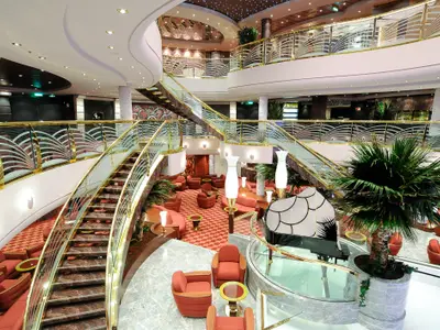 Das mehrstöckige Atrium der MSC Magnifica mit geschwungenen Treppen, goldenen Gelaendern, roten Moebeln und einem Piano, von oben fotografiert.