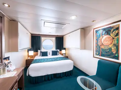 Aussenkabine an Bord der MSC Magnifica mit Doppelbett, runder Fensterluke und Blick auf das Meer. Links ein Schreibtisch, rechts ein Sofa und ein abstraktes Gemälde an der Wand.