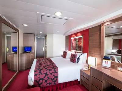 Eine helle und modern eingerichtete Balkonkabine an Bord der MSC Magnifica. Ein grosses Doppelbett mit roter Tagesdecke dominiert den Raum. Rechts ein Schreibtisch mit Spiegel und Steckdosen. Links ein TV und Kleiderschrank. Roter Teppichboden.