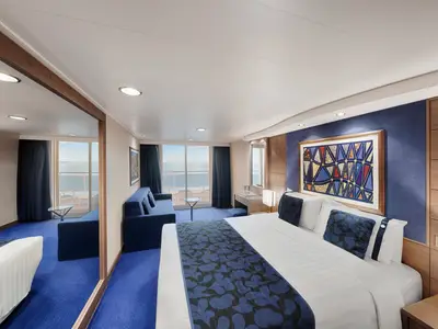 Eine moderne Balkonkabine an Bord der MSC Magnifica. Sie zeigt ein Doppelbett mit blauer Tagesdecke, ein blaues Sofa und einen Balkon mit Meerblick.