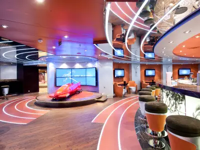 Die L'Olimpiade Bar an Bord der MSC Magnifica, mit einem roten Bobsled-Modell auf einer Buehne, einer langen Bar mit Hockern und mehreren Bildschirmen. Der Boden zeigt helle Linien eines Laufpfades.