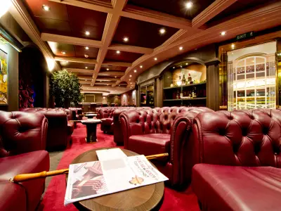 Die elegante Cuba Lounge Cigar Room an Bord der MSC Magnifica, ausgestattet mit zahlreichen roten Chesterfield-Ledersesseln, passendem roten Teppich und dunkler Holzvertäfelung. Ein Katalog liegt auf einem Tisch bereit.