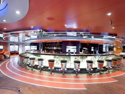 Moderne L'Olimpiade Sporting Bar an Bord der MSC Magnifica. Grosse Theke mit Barhockern, beleuchtete Laufbahn am Boden. Lounge-Bereiche sind sichtbar.