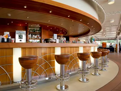 Einladende Bar-Lounge an Bord der MSC Magnifica mit einem eleganten, geschwungenen Tresen aus hellem Holz und mehreren Barhockern mit Lederbezug. Im Hintergrund ist die Küste zu sehen.