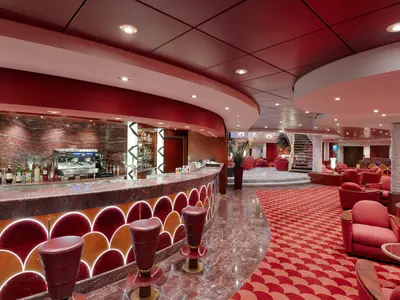 Die luxuriöse Le Gocce Bar an Bord der MSC Magnifica mit geschwungenem Marmortresen, dahinter eine Espressomaschine und Barregal. Roter Teppichboden und bequeme rote Loungesessel prägen den eleganten Raum.