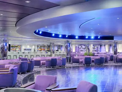 Moderne Onyx Lounge an Bord der MSC Magnifica. Blick auf lila Sofas, eine Bar und einen glänzenden Boden. Blaue und violette Deckenbeleuchtung schafft eine elegante Atmosphäre.