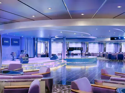 Die elegante Onyx Lounge an Bord der MSC Magnifica zeigt eine Bühne mit Schlagzeug, Grand Piano und viele violett-blaue Sitzbereiche sowie einen beleuchteten Boden.