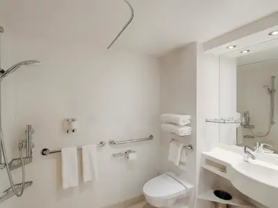 Ein modernes, barrierefreies Badezimmer an Bord der MSC Magnifica mit verstellbarer Dusche, Haltegriffen, Toilette und Waschtisch mit Spiegel. Weiße Handtücher liegen bereit.