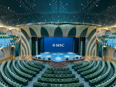 Weitwinkelaufnahme des Royal Theatre an Bord der MSC Magnifica mit leeren Sitzreihen, grosser Buehne mit MSC-Logo und sternenbesetzter Decke.