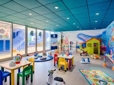Lebhafter Baby Club an Bord der MSC Magnifica. Der helle Raum bietet Rutschen, bunte Tische, Stühle und vielfältiges Spielzeug. Die Wände zeigen Unterwasser- und Strandmotive.
