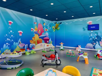 Bunter Kinderbereich an Bord der MSC Magnifica. Wände mit Meeresmotiven wie Fischen und Quallen. Viele Spielzeuge, darunter Babywippe, Motorrad, Schaukelpferd und Basketballkörbe, füllen den Raum.