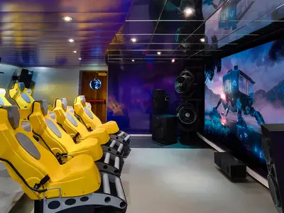 Innenansicht des 4D-Kinos an Bord der MSC Magnifica. Eine Reihe gelber, futuristisch anmutender Sitze ist auf eine grosse Leinwand ausgerichtet, die eine Szene mit einem roboterartigen Fahrzeug in einer fantastischen Landschaft zeigt. Seitlich sind Lautsprecher und Lüfter installiert.