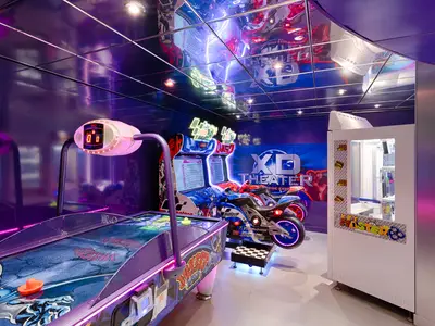 Bunte Spielhalle an Bord der MSC Magnifica mit Air Hockey Tisch, Motorrad-Rennsimulatoren und Greifautomat. Der Raum leuchtet in lila und blauen Neonlichtern.