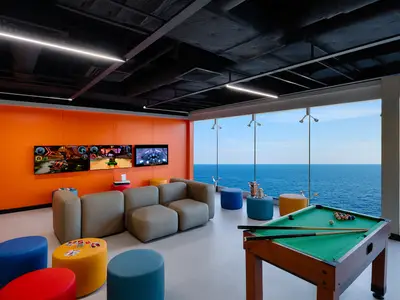Moderne Unterhaltungslounge im Young & Teens Club an Bord der MSC Magnifica. Orange Wände, Videospiele, Billardtisch und bunte Sitzgelegenheiten. Große Fenster bieten einen weiten Meerblick.