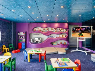 Ein farbenfroher Kinderbereich an Bord der MSC Magnifica mit Spieltischen, bunten Stühlen, einer Spielecke mit Konsolen und einem Bullauge zum Meer.