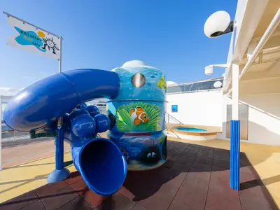 An Bord der MSC Magnifica: Bunter Kinderbereich mit blauer Röhrenrutsche und einem Ozean-Spielturm. Im Hintergrund sind ein Whirlpool und das weite Meer unter blauem Himmel zu sehen.
