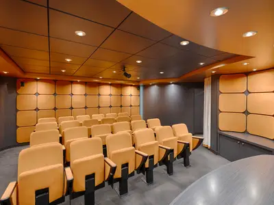 Moderner Konferenzraum an Bord der MSC Magnifica. Reihen von bequemen, orangefarbenen Polsterstühlen. Wände mit schalldämmenden Paneelen. Deckenleuchten und ein Projektor.