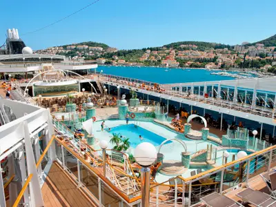 Panoramablick auf das belebte Sonnendeck der MSC Magnifica mit Pools, Liegestühlen, und Badegästen. Im Hintergrund die malerische Küste einer Stadt mit vielen Häusern.