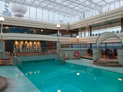 Ein helles, geräumiges Innenpool-Deck der MSC Magnifica, La Grotta Azzurra, mit türkisblauem Wasser, eleganter Fliesenarbeit und einem Glasdach. Seitliche Sitzbereiche und ein Rettungsring sind sichtbar.