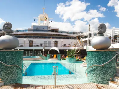 Das sonnige Pooldeck der MSC Magnifica mit Gästen im und am Wasser. Charakteristische Muschelskulpturen, türkis-grüne Mosaike und die Schiffsaufbauten prägen die Szene.