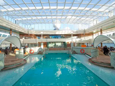 Innenpoolbereich La Grotta Azzurra an Bord der MSC Magnifica. Grosses Becken, helles Glasdach, Liegen und Dekorationen. Einige Personen sind entspannt zu sehen.