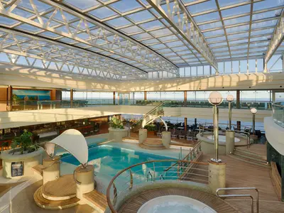 Blick auf die geräumige, überdachte Poollandschaft La Grotta Azzurra an Bord der MSC Magnifica, mit einem großen Pool, Whirlpools, Holzdecks, einer Bar im Hintergrund und weitem Meerblick durch die Fensterfront und das Glasdach.