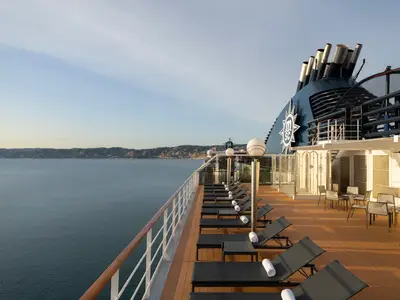 Ein weitreichendes Sonnendeck der MSC Magnifica mit zahlreichen dunklen Liegestühlen und weißen Handtüchern, flankiert von einem Geländer. Im Hintergrund das ruhige Meer und eine bergige Küstenlandschaft unter blauem Himmel. Der blau-weiße Schornstein des Schiffes ist rechts im Bild zu sehen.
