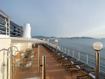 Weitläufiges Sonnendeck der MSC Magnifica mit Liegestühlen, Außenduschen und Sitzgelegenheiten. Im Hintergrund sieht man das Meer, eine Stadt und Berge unter hellem Himmel.
