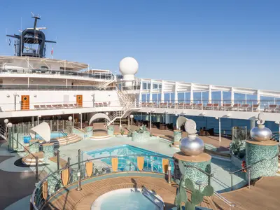 Weitwinkelansicht des Poolbereichs Le Grand Bleu auf der MSC Magnifica bei Tageslicht. Mehrere Pools und Whirlpools umgeben von Holzdecks und grünen Mosaiken. Zahlreiche Liegestühle und Sitzbereiche laden zum Entspannen ein, flankiert von dekorativen Skulpturen. Im Hintergrund ist das blaue Meer unter klarem Himmel sichtbar.
