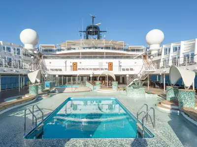 Panoramablick auf den Le Grand Bleu Poolbereich an Bord der MSC Magnifica. Mehrere Pools und Whirlpools, umgeben von Liegestühlen und einer Bar, unter blauem Himmel. Keine Personen sichtbar.