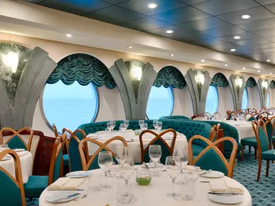 Das elegante L'Edera Restaurant an Bord der MSC Magnifica zeigt stilvoll gedeckte Tische, türkisfarbene Sofas und Holzstühle. Runde Bullaugenfenster geben den Blick auf das weite Meer frei.