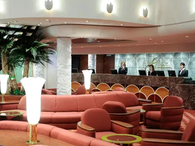 Die Rezeption 'Le Gocce' der MSC Magnifica mit drei Mitarbeiterinnen hinter einem Marmortresen, umgeben von einer Lounge mit roten Sofas, Sesseln und dekorativen Palmen.