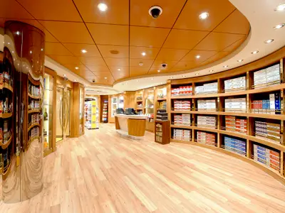 Modern duty-free shop onboard the MSC Magnifica. Light wooden shelves stock tobacco and snacks. A central counter and reflective pillar stand out.