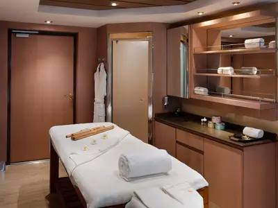 Ein luxuriöses Behandlungszimmer im MSC Aurea Spa an Bord der MSC Magnifica. Eine weisse Massageliege steht im Vordergrund, dahinter ein Waschtisch mit Pflegeprodukten und ein Spiegelregal mit gerollten Handtüchern.