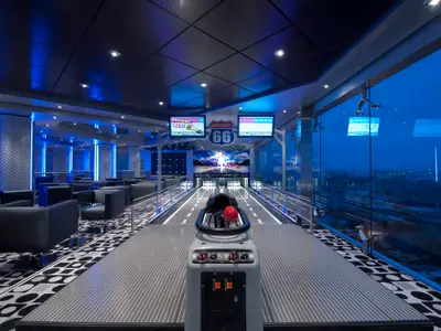 Weitwinkelaufnahme der modernen Bowlingbahn 'Highway 66' und Lounge an Bord der MSC Magnifica. Blaue LED-Lichter beleuchten den Raum, grosse Fenster bieten eine Aussicht auf die blaue Stunde.