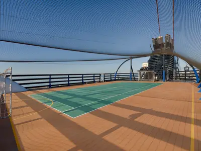 Multifunktions-Sportplatz an Bord der MSC Magnifica auf dem Oberdeck bei Tageslicht. Der grüne Platz mit Toren und Basketballkorb ist von einem Schutznetz umgeben und leer.