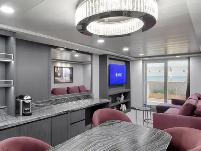 Eine moderne Suite an Bord der MSC Magnifica mit einem eleganten Wohnzimmer, gemütlichem Sofa und einem Balkon mit Meerblick. Dunkle Holzmöbel, Marmoroberflächen und ein großer TV sind sichtbar.