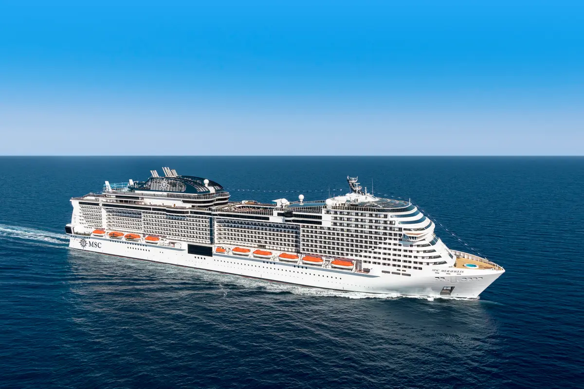 Die MSC Meraviglia auf dem offenen Meer unter blauem Himmel, aus der Vogelperspektive. Das grosse weisse Kreuzfahrtschiff hinterlaesst eine weisse Welle auf dem blauen Wasser.