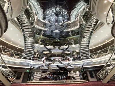 Ein luxuriöses, mehrstöckiges Atrium an Bord der MSC Meraviglia mit geschwungenen Spiegeltreppen, einem Grand Piano und einer glänzenden, reflektierenden Decke, die den Raum optisch erweitert.