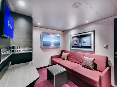 Heller Wohnbereich einer Aussenkabine an Bord der MSC Meraviglia. Rotes Sofa, Beistelltisch, TV und ein bullaugenartiges Fenster mit Meerblick sind zu sehen. Die moderne Kabine ist einladend gestaltet.