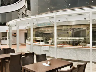 Die Atmosphere Bar South und das Marketplace Buffet an Bord der MSC Meraviglia. Ein heller Bereich mit Esstischen, Stühlen und einem Buffet mit verschiedenen Speisen hinter Glas.
