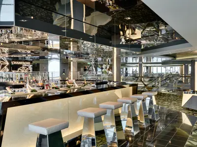 Blick in die moderne Champagne Bar an Bord der MSC Meraviglia. Eine lange Bar mit Champagnerflaschen und Gläsern, davor spiegelnde Barhocker. Die Decke reflektiert den Raum, im Hintergrund weitere Ebenen des Schiffes.