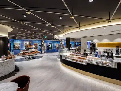 Blick in das stilvolle Jean-Philippe Chocolat & Café an Bord der MSC Meraviglia. Ein langer Tresen mit Suessigkeiten und Getraenken, umgeben von eleganten Shops und einer modernen Deckenbeleuchtung.